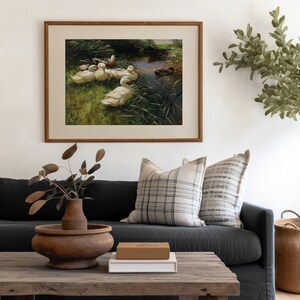 May include: A framed painting of a group of white ducks swimming in a pond with green reeds. The painting is in a wooden frame and is hanging on a white wall.