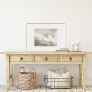 May include: A light wood console table with three drawers and two baskets. The table has a framed black and white print of clouds above it. The print is in a gold frame. There are two white candlesticks on the table with white candles.
