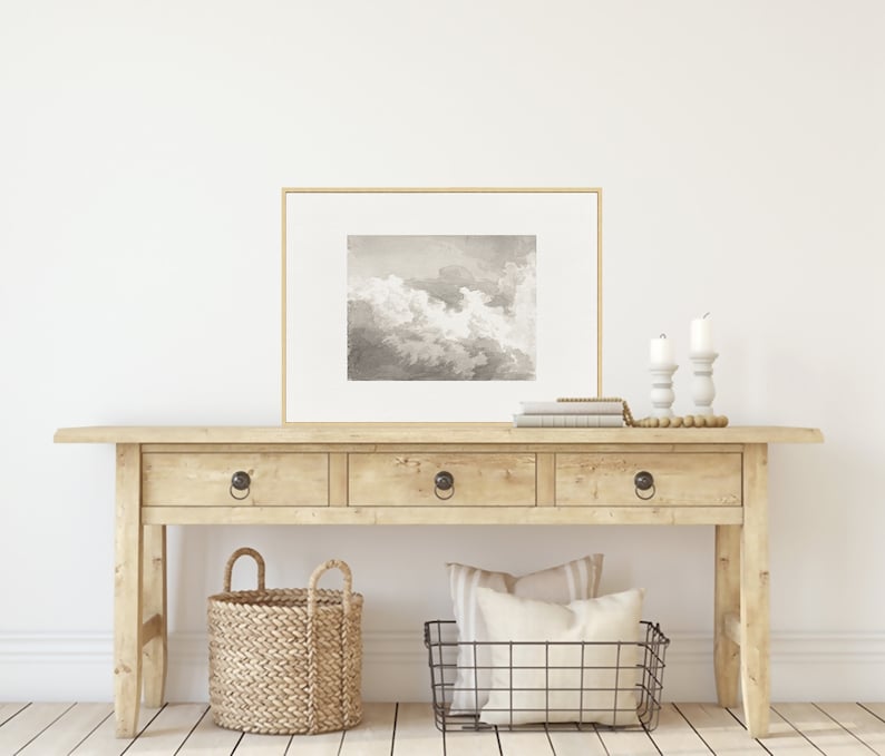 May include: A light wood console table with three drawers and two baskets. The table has a framed black and white print of clouds above it. The print is in a gold frame. There are two white candlesticks on the table with white candles.