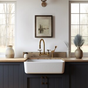 May include: A white farmhouse kitchen sink with a gold faucet and a wooden countertop. The sink is in front of a window with a view of trees. There is a framed picture of sailboats on the wall above the sink.