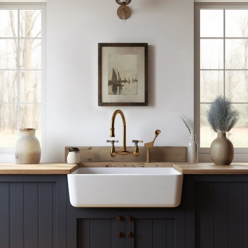 May include: A white farmhouse kitchen sink with a gold faucet and a wooden countertop. The sink is in front of a window with a view of trees. There is a framed picture of sailboats on the wall above the sink.