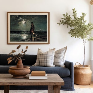 May include: A framed painting depicting a figure walking along a beach at night, holding a lantern. The painting is hung on a white wall above a dark blue sofa with cushions. A potted plant stands in the corner of the room.