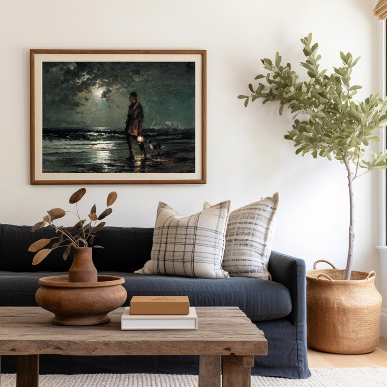 May include: A framed painting depicting a figure walking along a beach at night, holding a lantern. The painting is hung on a white wall above a dark blue sofa with cushions. A potted plant stands in the corner of the room.
