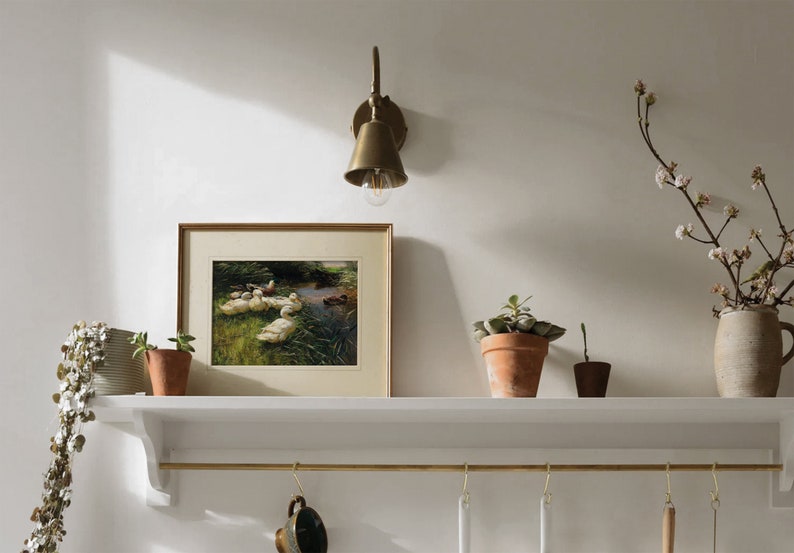 May include: A white shelf with a brass rail and hooks hangs on a white wall. A framed print of ducks swimming in a pond sits on the shelf. There are three potted plants on the shelf, and a brass wall sconce with a light bulb is mounted on the wall above the shelf.