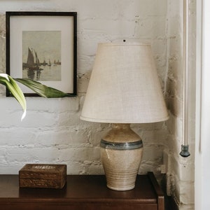 May include: A ceramic table lamp with a beige and blue patterned base and a white linen lampshade. The lamp is sitting on a dark brown wooden surface.