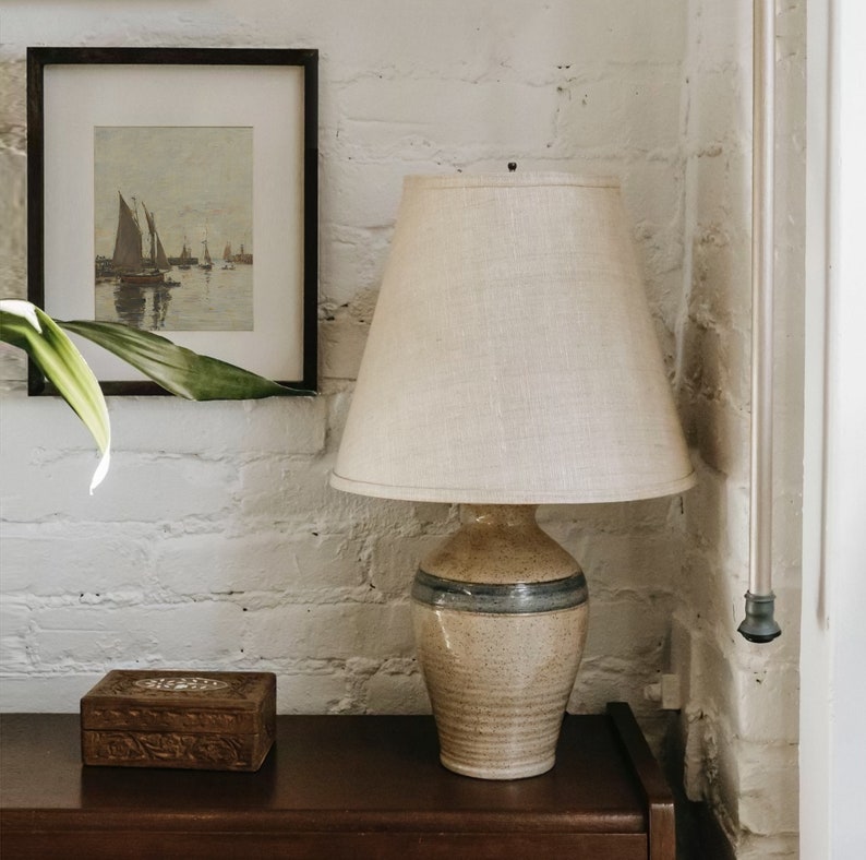 May include: A ceramic table lamp with a beige and blue patterned base and a white linen lampshade. The lamp is sitting on a dark brown wooden surface.