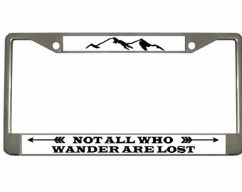 May include: A silver metal license plate frame with a white background and black text and a mountain silhouette. The text reads "Not all who wander are lost" with an arrow pointing to the right and an arrow pointing to the left.