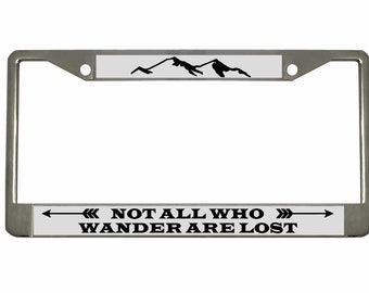 May include: A silver metal license plate frame with a white background and black text and graphic. The text reads "Not all who wander are lost" with arrows on either side. The graphic is a silhouette of mountains.