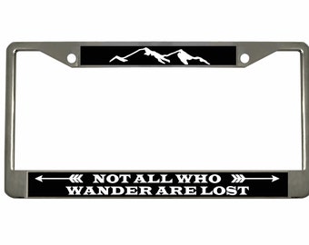 May include: A silver license plate frame with a black background and white text that reads "NOT ALL WHO WANDER ARE LOST". The top of the frame has a white mountain silhouette on a black background.