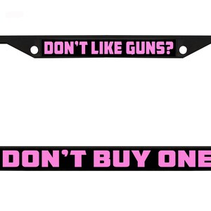 May include: Black license plate frame with the text "DON'T LIKE GUNS? DON'T BUY ONE" in bright pink lettering. The frame is rectangular with rounded corners and a space for the license plate.