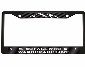 May include: Black metal license plate frame with a carbon fiber pattern and white text that reads "NOT ALL WHO WANDER ARE LOST" with a white mountain range graphic and white arrows on either side of the text.