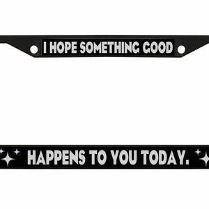 May include: Black license plate frame with white text that reads "I HOPE SOMETHING GOOD HAPPENS TO YOU TODAY." The frame has a rectangular shape with rounded corners and a black border.