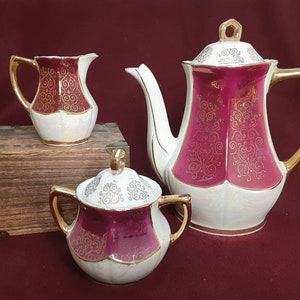 Puede incluir: Un juego de té blanco con ribetes dorados y un diseño floral burdeos. El juego incluye una tetera, una jarra de crema y un azucarero.
