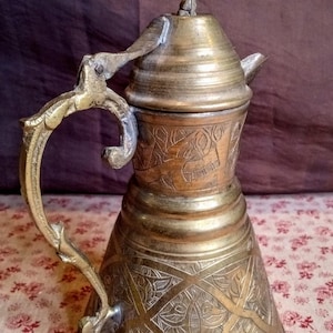 Puede incluir: Una cafetera antigua de latón con intrincados diseños grabados. La cafetera presenta un asa larga y curvada y una tapa decorativa con un remate único. El cuerpo de la cafetera tiene forma bulbosa, y el color general es una mezcla de oro y bronce.