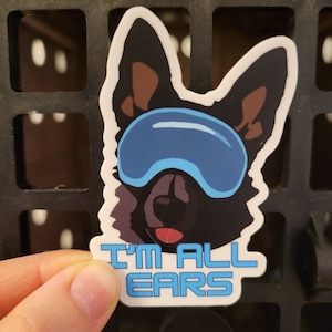 May include: A white sticker with a black and brown cartoon dog wearing blue goggles. The text "I'm All Ears" is printed in blue on the sticker.