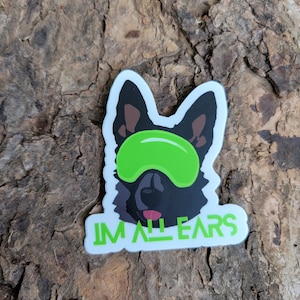 May include: A black and white sticker of a dog's head with green eyes and a green headband. The text "I'M ALL EARS" is written below the dog's head.