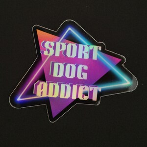 May include: A sticker with the text "SPORT DOG ADDICT" in a pixelated font. The text is inside a neon-outlined triangle with a gradient of orange, pink, and purple. The background is black.