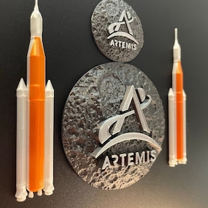 May include: Three 3D printed models of the Space Launch System (SLS) rocket, one orange and two white, are displayed next to two silver metal Artemis logos. The logos are embossed with the Artemis logo and the text "ARTEMIS".