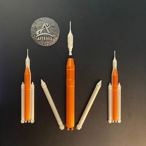 Peut inclure: Un ensemble de cinq fusées modèles blanches et orange. Les fusées sont disposées en ligne sur une surface noire. Les fusées sont de tailles différentes, la plus grande étant au centre. Les fusées sont toutes en plastique et ont un design réaliste.