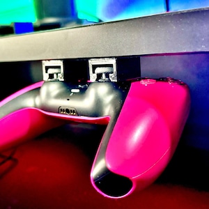 Puede incluir: Un mando de videojuego rosa y negro está montado en un estante negro con un soporte negro. El mando está colgado boca abajo.