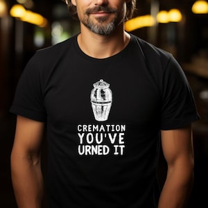 以下が含まれることがあります： 白い壷と「CREMATION YOU'VEURNED IT」という文字が書かれた黒いTシャツ。