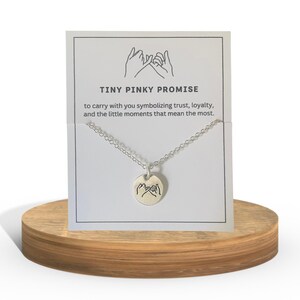 May include: A silver necklace with a round pendant featuring a pinky promise design. The necklace is displayed on a wooden stand with a card that reads "TINY PINKY PROMISE" and a description about trust and loyalty.
