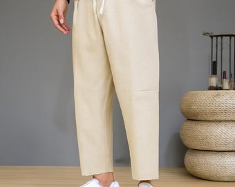 May include: Beige linen pants with a drawstring waist and white sneakers. The pants are a neutral color and have a relaxed fit. The outfit is completed with a white t-shirt.