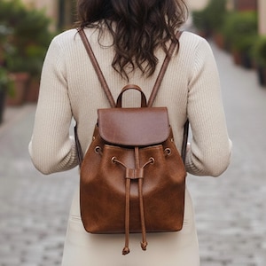 May include: A brown leather backpack with a flap and drawstring closure. The backpack has adjustable straps and a top handle. The bag is worn on the back of a person wearing a cream-coloured sweater and trousers. The background is a cobbled street.