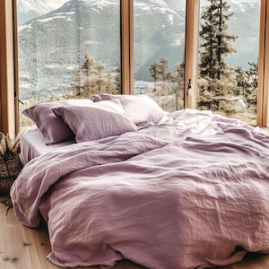 Op de afbeelding: Een bed met lavendelkleurig linnen beddengoed, inclusief een dekbed en kussens, staat voor een groot raam met uitzicht op de bergen. Een gevlochten mand met gedroogde planten staat op de vloer. Een rotan stoel staat op de achtergrond.