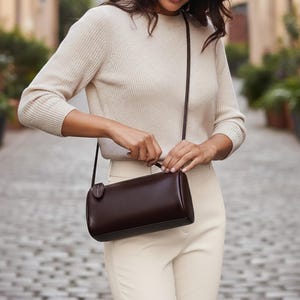 Peut inclure: Un sac bandoulière cylindrique marron foncé avec une longue et fine bandoulière. Le sac est en cuir lisse et comporte une petite étiquette circulaire attachée. La personne porte un pull et un pantalon crème.