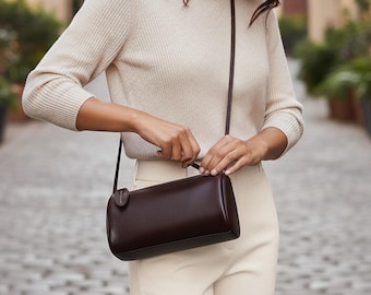 Puede incluir: Un bolso bandolera cilíndrico marrón oscuro con una correa larga y fina. El bolso es de cuero liso y tiene una pequeña etiqueta circular adjunta. La persona lleva un jersey y unos pantalones color crema.