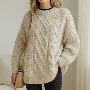 Puede incluir: Un suéter beige de punto trenzado con cuello redondo y mangas largas. El suéter se combina con vaqueros negros y botines color canela. Un bolso de mano beige con correa de cadena se lleva al hombro. El conjunto es informal.