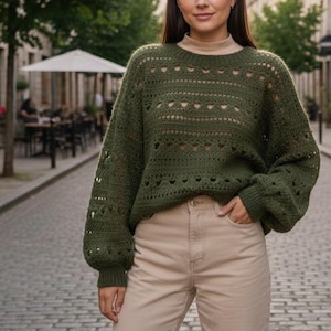 May include: An olive green knitted jumper with heart-shaped cut-outs and long sleeves. The jumper is paired with beige trousers. The person is standing on a cobbled street.