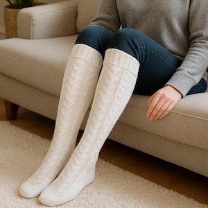 May include: Cream-colored cable knit over-the-knee socks. The socks have a ribbed cuff and a textured cable knit pattern. The person in the image is wearing the socks with blue jeans and a gray sweater. The socks are made of a soft, warm material.