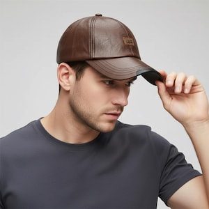 Puede incluir: Gorra de béisbol de cuero marrón con visera curva y un pequeño parche rectangular en la parte delantera. La gorra es de color marrón liso y tiene un diseño estructurado. La visera es negra por debajo.