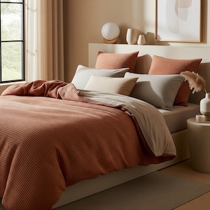 Puede incluir: Una cama con ropa de cama de tejido de gofre de color terracota, que incluye un edredón y almohadas a juego. Almohadas adicionales en gris claro y beige proporcionan contraste. La escena está ambientada en un dormitorio de tonos neutros con una ventana y detalles decorativos.