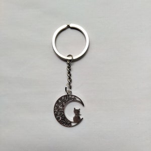 May include: Silver keychain featuring a crescent moon with an intricate design and a silhouette of a cat sitting on the moon.