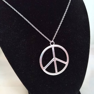 May include: A silver peace sign pendant necklace on a silver chain. The necklace is displayed on a black velvet stand.