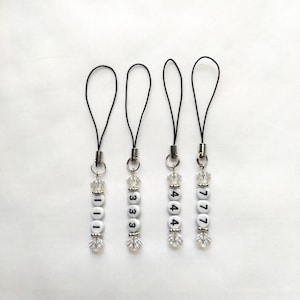 May include: Four beaded phone charms with black cords. Each charm has white letter beads spelling out numbers: 1111, 3333, 4444, and 7777. Clear, faceted beads are interspersed between the letter beads.