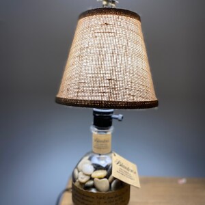 Puede incluir: Una lámpara de mesa hecha con una botella de whisky reutilizada con una pantalla de lámpara de arpillera. La botella está llena de piedras pequeñas y tiene una etiqueta que dice "Wilderness".