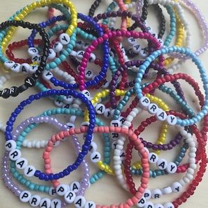 May include: A collection of colorful beaded bracelets. Each bracelet is made of small, round beads in various colors, including pink, blue, yellow, and black. Some bracelets spell out the word "PRAY" with white letter beads. The bracelets are scattered.