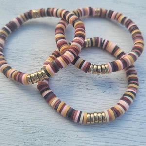 May include: Three colorful bracelets made of stacked, round beads in shades of brown, pink, black, and yellow. Each bracelet has a gold metal accent.