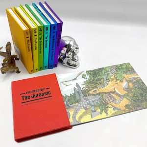 以下が含まれることがあります： 「THE MESOZOIC - The Jurassic」というタイトルの赤い本が、カラフルな本の積み重ねの後ろにあります。本は、ジャングルの設定で恐竜のイラストが描かれたページを開いています。