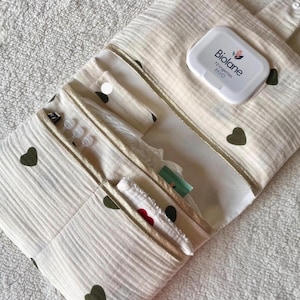 Può includere: Un organizer in tessuto color crema con un motivo a cuori verde scuro e bordi dorati. L'organizer presenta diverse tasche interne e un dispenser di salviette in plastica bianca Biolane 72 lingettes H2O fissato alla patta con un bottone.
