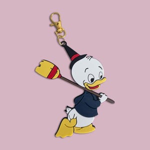May include: A rubber keychain featuring a cartoon duck character wearing a dark blue jacket and a black pointed hat with a red band. The character holds a yellow broom with a red handle over its shoulder. The keychain has a gold-colored metal clasp.