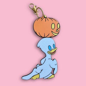 May include: A keychain featuring a smiling orange pumpkin and a light blue cartoon duck. The pumpkin has black outlines and a gold-colored clasp. The duck has blue eyes and yellow feet. The background is a solid pink color.