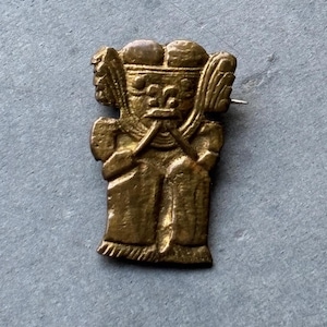 以下が含まれることがあります： ヘルメットと腕を上げた様式化された人物が特徴の、ヴィンテージの金色の金属ピン。 この人物は詳細な顔をしており、長い衣服を着ています。 ピンにはシンプルなピンバックが付いています。