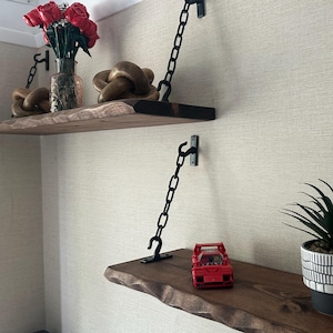 May include: Two rustic wooden shelves with live edges are mounted on a wall with black chain supports. The top shelf holds a vase of red roses and wooden decorative knots. The bottom shelf displays a red toy car and a potted plant.
