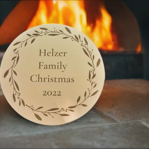 May include: A gold-colored round ornament with a wreath design and the text "Helzer Family Christmas 2022".
