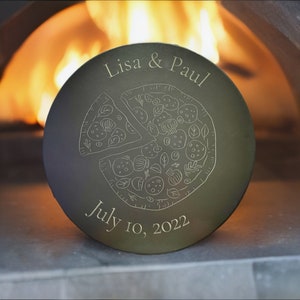 May include: A round, dark gray slate pizza serving board with an engraved design of a pizza with toppings and the text "Lisa & Paul" and "July 10, 2022".
